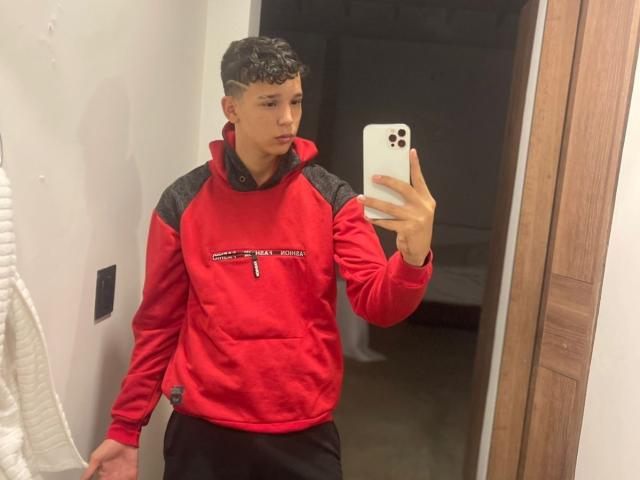 AxelBiel, short curly black hair, red and gray hoodie, black pants, holding white iPhone, taking mirror selfie, medium build, light skin, serious expression, standing in dimly lit room.