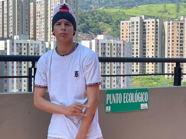 AxelBiel wears a white tee, red and black cap, and white shorts, standing with a slight smirk, hands near his crotch, in a city rooftop setting.