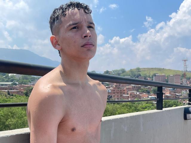 AxelBiel, shirtless, sweaty, with curly black hair, standing on a rooftop, looking up, blue sky with clouds, green hills, and urban buildings in the background.