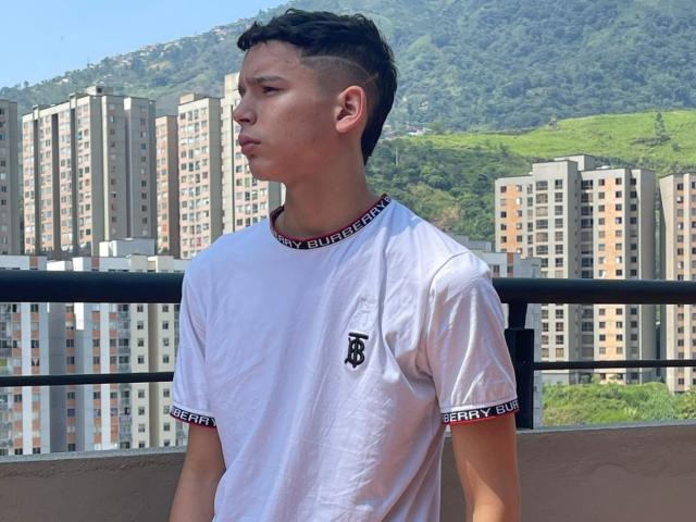 AxelBiel with a white t-shirt, "B" logo, and "VERY B" cuffs, standing on a balcony, looking to the side, short dark hair, slight tan, mountain and buildings in background.