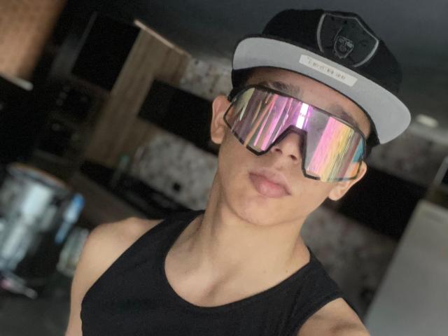 AxelBiel with reflective sunglasses, black tank top, and black/gray cap, sticking out tongue, showing a muscular, cute face with medium skin and short hair.