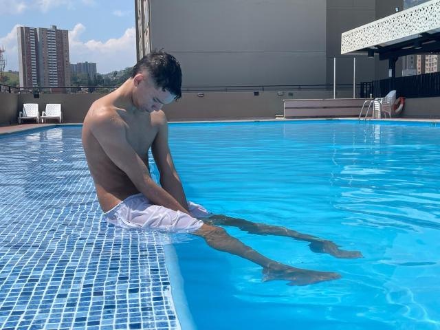 AxelBiel, shirtless, with white swim trunks, sitting in a blue-tiled pool, water up to his thighs, hands on pool edge, looking down, short black hair, urban rooftop.