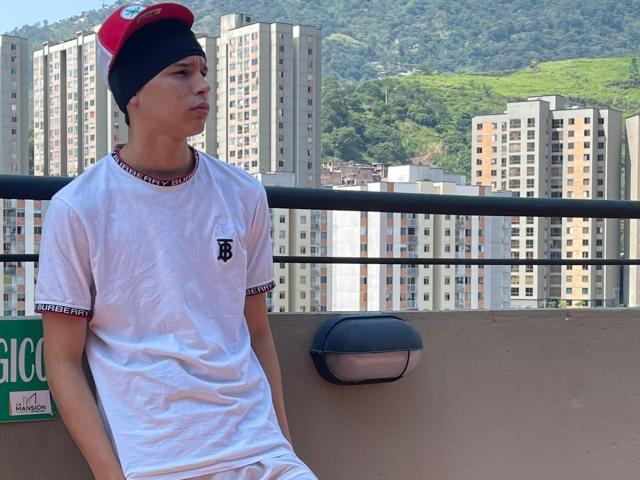 AxelBiel, wearing a red and black cap, white tee with a black "B" and black trim, sitting on a balcony, looking to the side, with a wall light.