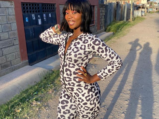 EbonyAngel-hot in a leopard-print jumpsuit, standing on a dirt road, hand on hip, smiling, dark skin, black hair, and a ring on her finger. Shadows on the ground.