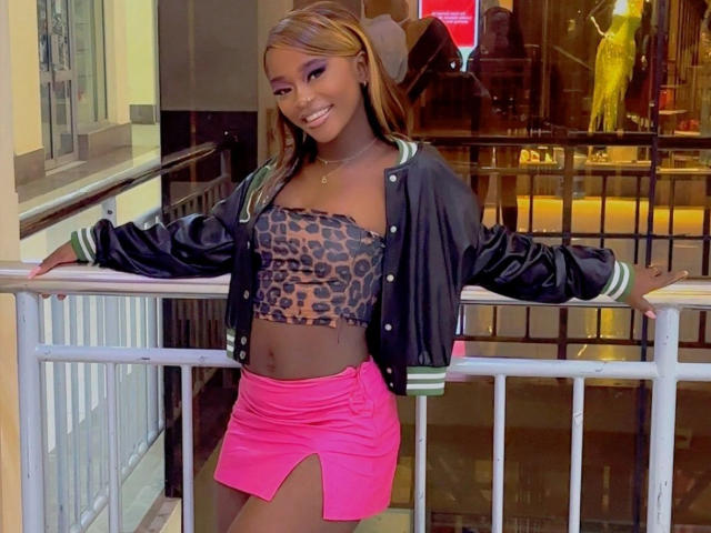 EbonyAngel-hot with dark skin, smiling, wearing a leopard print tube top, black jacket, and pink skirt with a side slit, standing against a railing, arms out.