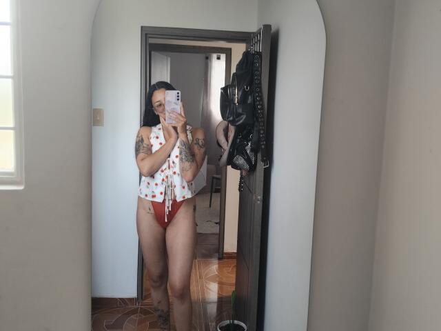 A mirror selfie of Mllevv standing in a hallway. Mllevv has dark skin, long straight black hair, and several tattoos on her arms. She wears a white polka dot crop top with red high-waist panties, showing her full, curvy figure. She holds a phone with a pink case in her right hand. A black leather jacket hangs on the right wall. The floor has wooden patterns. Natural light from a window on the left.