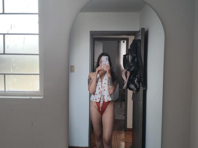 Mllevv stands in front of a mirror, wearing a red thong and a white shirt with red patterns, showing tattoos, holding a phone, with boots hanging on the door.