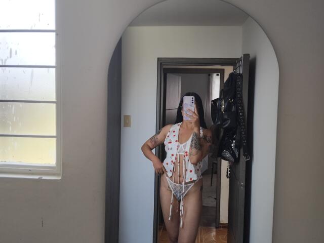 Mllevv stands in a mirror, wearing a white lace lingerie set with red heart patterns. They have tattoos on their left arm and right side. Mllevv's face is partially covered by a phone. Mllevv's hair is dark, and they are holding a black leather belt in their right hand. The mirror is framed in a white arch, with a window to the left and a doorway behind Mllevv. The room has wooden floors. The image is a casual selfie with a focus on Mllevv's lingerie and tattoos.