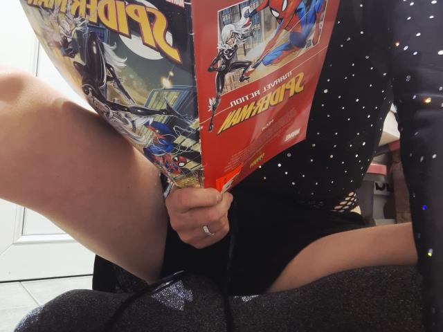 Millfka's legs spread, holding a Marvel comic, wearing a black, sparkly dress, with a ring on her finger, sitting on a gray, glittery surface.