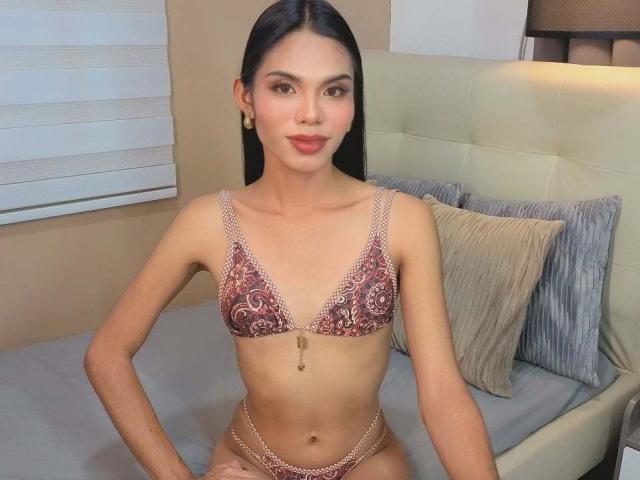 CassandraWise with long black hair, wearing a red and pink patterned bra and matching panties, sitting on a bed with hands on her hips, looking at the camera.