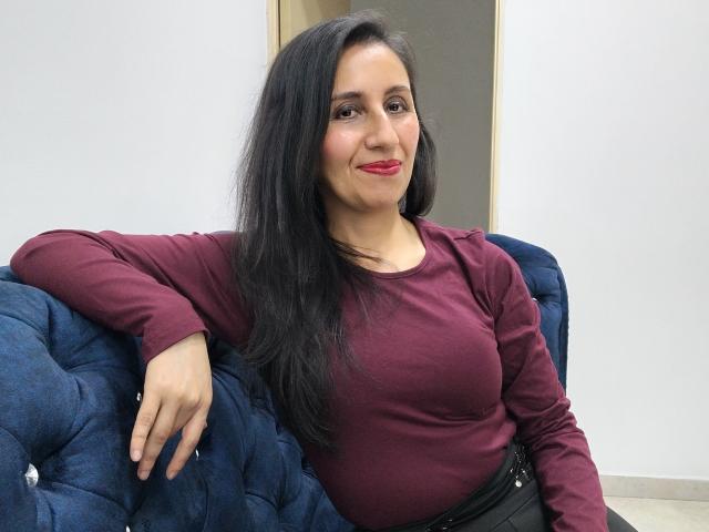 Cyntiia with long black hair, red lipstick, and a burgundy top, sitting on a blue tufted couch, smiling, hand on her thigh, black belt, casual look.