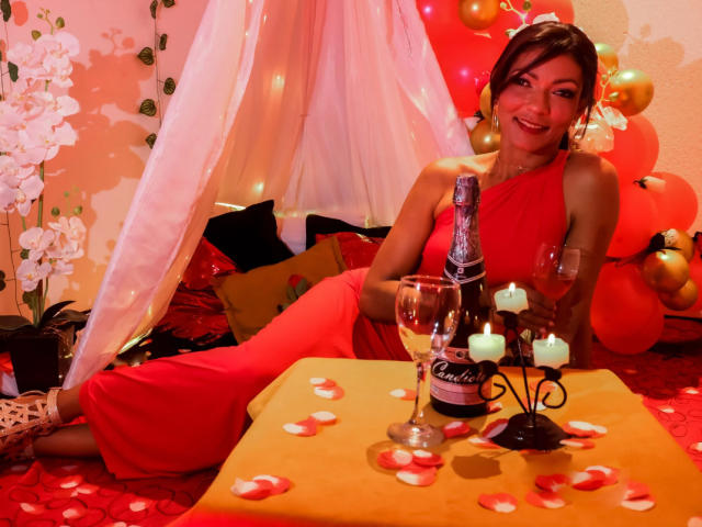 HannaPretty in a red one-shoulder dress, sitting on a red carpet, holding a champagne bottle, with candles and rose petals on a yellow table, smiling.