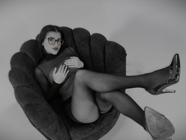 Iris Blair mit Brille, in einem dunklen Kleid, sitzend in einem bequemen Sessel, mit verschränkten Beinen, wobei ihr Hintern zu sehen ist, in schwarzen High Heels, die ihre Brüste haltend, auf einem Schwarz-Weiß-Foto..