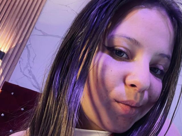 GraceRedMiller, con i capelli castano striati di viola, gli occhi azzurri e un sorriso appena accennato, con le labbra leggermente corrugate. Il suo naso è centrato e indossa una maglietta bianca..
