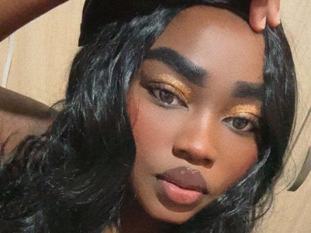 ReinaEbanox has dark brown skin, thick black eyebrows, gold eyeshadow, and full pink lips. Her black wavy hair is partially covered by a black hat.