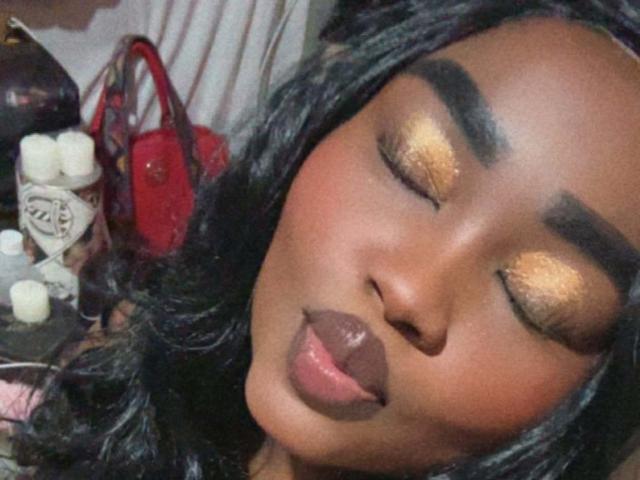 ReinaEbanox's face with closed eyes, golden shimmer eyeshadow, thick eyebrows, dark lipstick, black wavy hair, red purse in background, white candles, and red bag on the left.