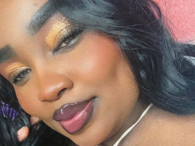 ReinaEbanox's face with shiny gold eyeshadow, dark brown eyes, glossy pink lips, and black wavy hair, looking slightly to the side with a subtle smile. Her skin is dark.