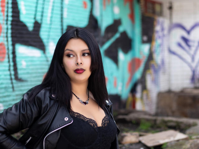 GabyJoy with black lace bra, black leather jacket, and red lipstick, standing with hands on hips, has a necklace, and medium tits, with long black hair, looking serious.