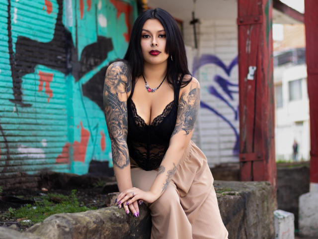 GabyJoy with tattoos, black lace top, big tits, tan pants, leaning on a stone wall, black hair, red lipstick, pink necklace, looking at camera, graffiti behind.