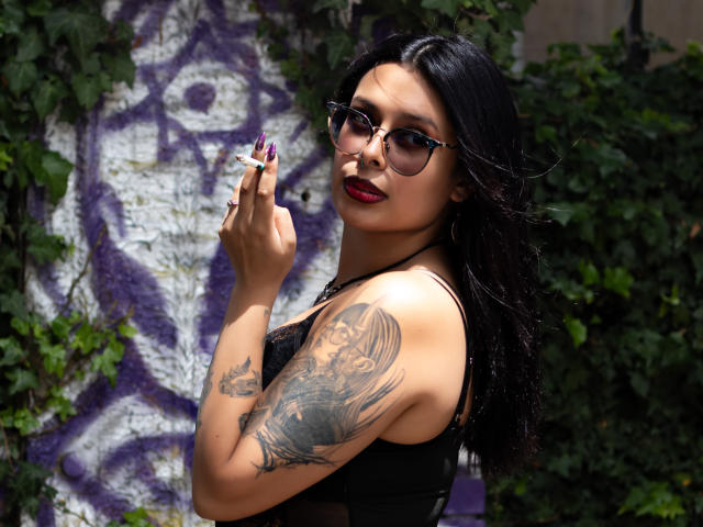 GabyJoy with black hair, glasses, red lipstick, holding cigarette, showing tattooed arm, wearing black top, standing in front of graffiti-covered wall with green vines.