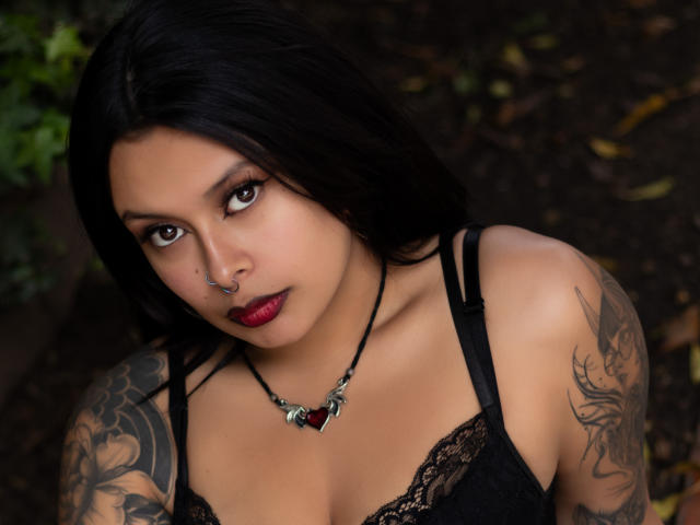 GabyJoy with dark hair, red lipstick, black lace top, tattoos on shoulders, nose ring, and black necklace with red heart, looking up with serious eyes.