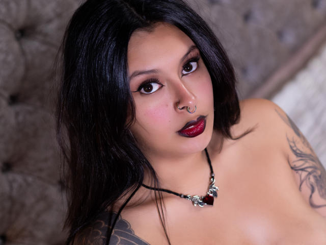 GabyJoy with dark hair, nose ring, and red lipstick, looking up with dark eyes, wearing a black necklace with a red gem, and showing tattoos on her upper arm.