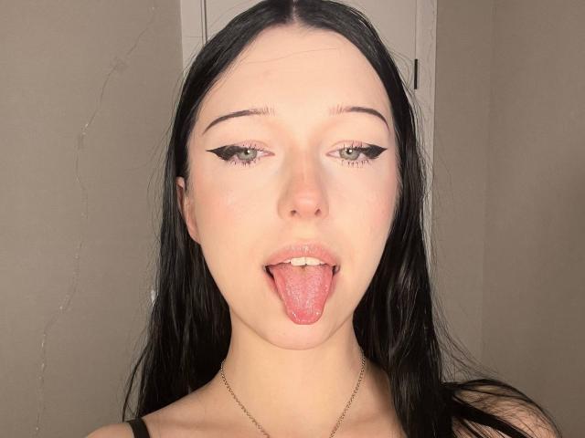 AmaiRin has pale skin, long black hair, green eyes, and is sticking out her tongue with black eyeliner, wearing a thin necklace. Boobs not visible, pussy not visible.