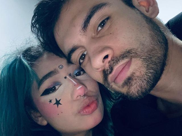 NightControl with teal hair and makeup, nose ring, black star face paint, and black eyeliner, leans on a bearded NightControl with a nose piercing, looking into the camera.