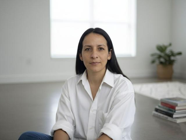 CharlotteAshford is a medium-skinned woman with straight, black hair sitting on a gray floor. She wears a white button-up shirt and blue jeans. Her expression is neutral, with a slight smile. She's centered in the image, with a stack of books to her right and a potted plant in the background. The bright window behind her creates a soft, natural light. The composition is simple and focused on CharlotteAshford.