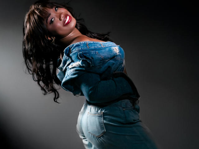 KarlieMoss with long dark hair, red lipstick, and blue ripped jeans, smiling, looking back, showing her ass, hands on chest, dark background, studio light, sexy, confident.