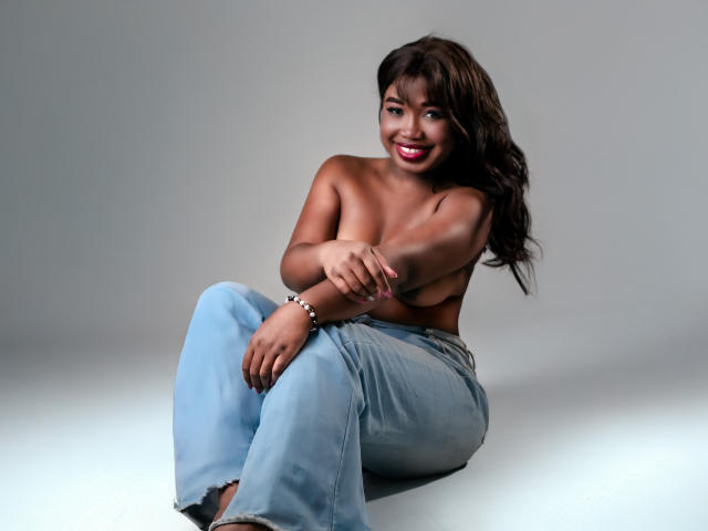KarlieMoss with dark brown hair, topless, wearing light blue jeans, sitting with knees up, arms crossed over small tits, smiling with red lipstick, gray background, looking at camera.