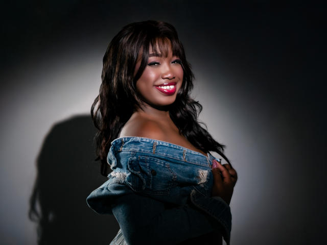 KarlieMoss with dark skin, wavy black hair, and red lipstick, smiling, wearing a denim off-shoulder shirt, showing her small, perky tits, in a dark studio.