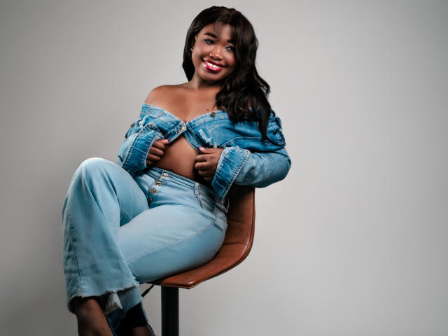 KarlieMoss with dark skin, long wavy black hair, red lipstick, and a smile, wearing an off-shoulder denim jacket and light blue jeans, sitting on a brown chair.
