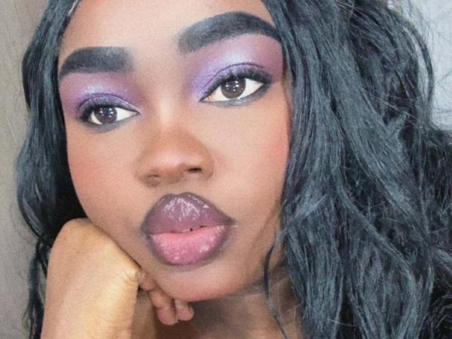 ReinaEbanox has dark purple eyeshadow, glossy purple lips, and black wavy hair, with her hand under her chin, looking up with brown eyes, and thick, dark eyebrows.