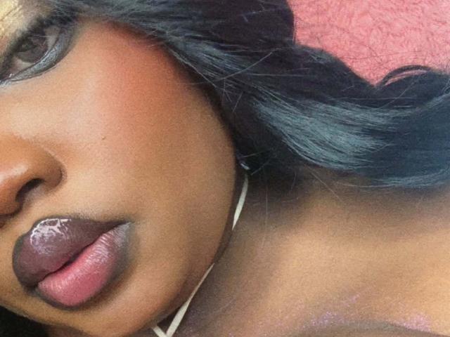 ReinaEbanox's dark brown face, glossy black hair, and shiny purple lipstick. Her soft, brown skin and slightly parted lips are in focus. She's wearing a thin white strap.