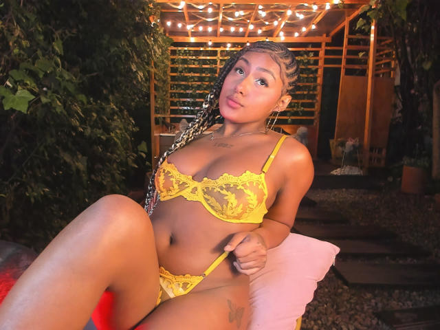 AmberQueeny in yellow lace lingerie, sitting with legs apart, showing her tits and pussy, braided hair, light brown skin, cute face, outdoor patio, string lights in background.