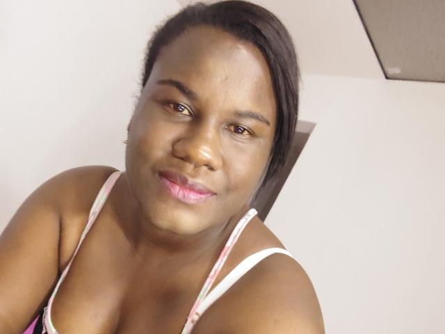 Close-up photo of KartHemos. KartHemos has dark brown skin, short black hair pulled to the side, and a slight smile. She has pink lipstick and wears a floral-patterned spaghetti strap top. Her eyes are slightly squinted. The lighting is even, highlighting her smooth skin. The upper part of a white wall and a brown rectangular shape are in the background. The image has a casual, informal feel.