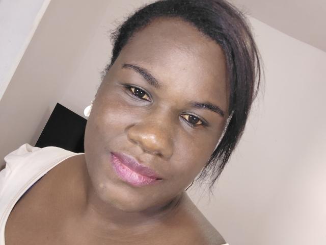 Close-up of KartHemos, a dark-skinned woman with medium-length black hair in a messy updo. She has brown eyes, subtle eyeliner, and pink glittery lipstick. KartHemos wears a white off-shoulder top, revealing her neck. Her skin is smooth with a slight sheen. The background is plain and out of focus. The image has a casual, slightly tilted angle, emphasizing KartHemos' relaxed expression. The lighting is even, highlighting her facial features.