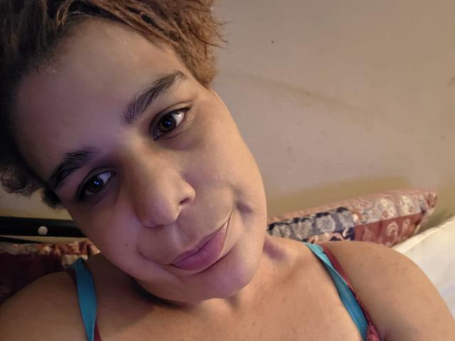 CockTease with short curly brown hair, brown eyes, and light brown skin, sticking out her tongue and wearing a blue tank top, showing her small tits.