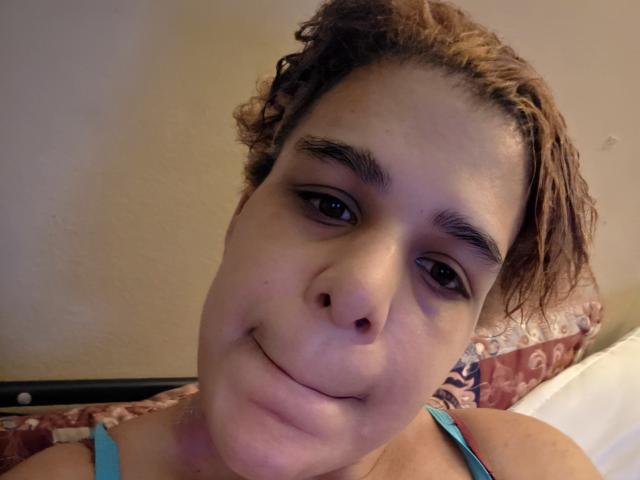 CockTease has light brown skin, curly brown hair, and is smiling with a slight pout. She has thick eyebrows, brown eyes, and is wearing a blue top.