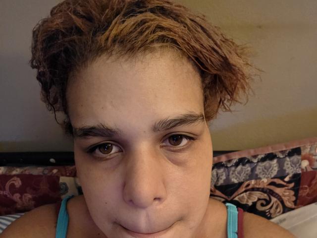 Close-up of CockTease with curly, light brown hair, fair skin, and brown eyes. CockTease is looking directly at the camera with a slightly pouty expression, lips pressed against the bottom of the chin. She has dark eyebrows and wears a light blue tank top with thin straps. The background shows a patterned pillow with red, blue, and beige floral designs. The image has a casual, unfiltered feel, with natural lighting and slight shadow on the right side of the face. The texture of the hair is slightly curly and unkempt.