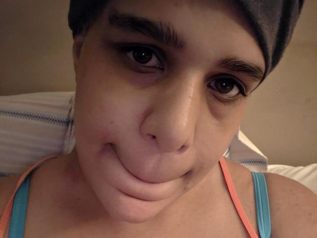 Close-up of CockTease with a short, dark-haired cap, brown eyes, and a subtle smile. CockTease has a round face, slightly parted lips, and a small pimple on the left cheek. Wearing a teal tank top with orange straps. The image is focused on CockTease's face, showing clear skin and dark eyebrows. Soft, warm lighting highlights the facial features. The background is blurred. The overall composition is intimate and direct.