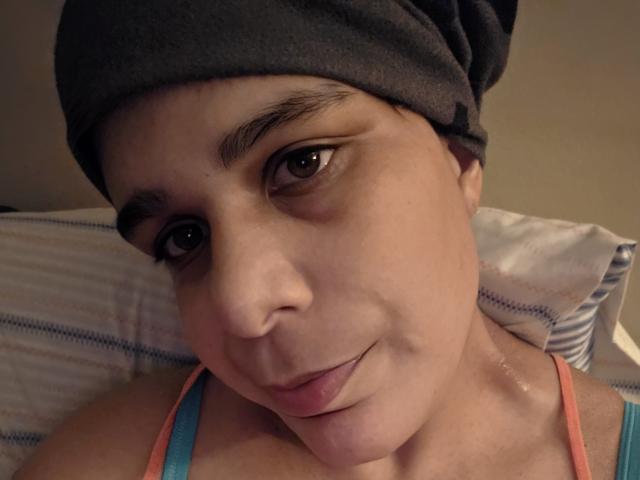CockTease with light brown skin, dark eyebrows, and brown eyes, wearing a gray headband, and pink and blue tank top, sticking out her tongue slightly.