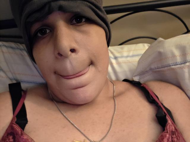 CockTease has a beanie on, tongue slightly out, wearing a red lace bra, showing soft tits with black straps, and a silver necklace, lying on a bed.