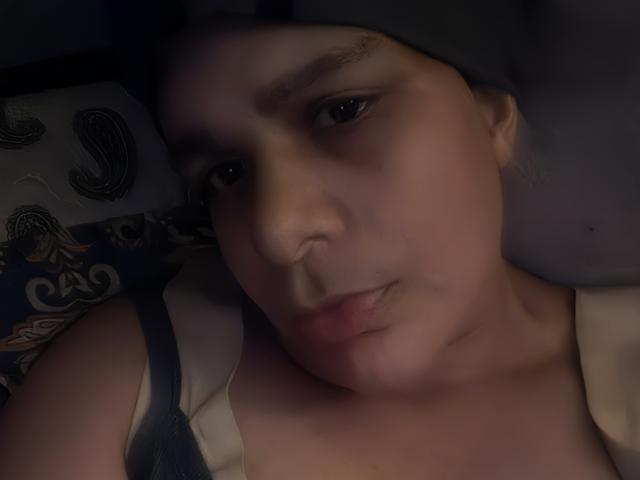 A dimly lit selfie of CockTease with a slightly pouty expression, wearing a tank top and a gray headscarf, showing a small piercing on the lower lip.