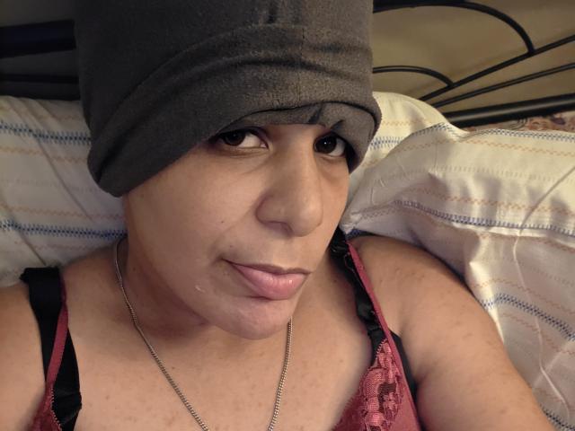 Close-up photo of CockTease lying on a bed. CockTease has light brown skin, small nose, and slightly parted lips. She wears a gray beanie and a red lace bra with black straps. Her brown eyes look directly at the camera. A silver chain necklace rests around her neck. The bed has a striped pillow with white and blue lines. The metal headboard's black pattern is partially visible. The lighting is soft, highlighting her facial features. The composition focuses on CockTease's upper body and face.