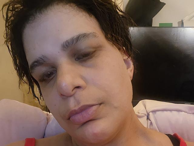 Close-up photo of CockTease, a light-skinned woman with wet, curly dark brown hair, looking slightly downward with a subtle pout. She has small, dark eyebrows, slightly droopy eyelids, and full, pinkish lips. She's wearing a red and black top. The background is blurred, showing a black headboard and a pink pillow. The image focuses on her facial expression and texture of her skin, highlighting her natural, slightly messy look. Her eyes are small and slightly asymmetrical. The lighting is soft and even.