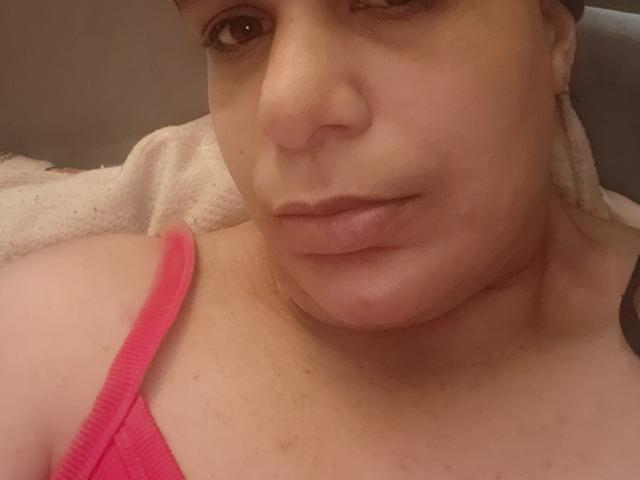 Close-up of CockTease's face, showing a light skin tone with a slightly flushed complexion. CockTease has medium brown eyes, a small nose, and full lips with a neutral expression. They are wearing a pink ribbed tank top with a thin strap, visible on the left side of the image. The background is blurry, focusing on CockTease's upper chest and face. The image has a soft, warm light. Slight texture on the skin, with some small blemishes visible. Overall, a straightforward, intimate portrait.