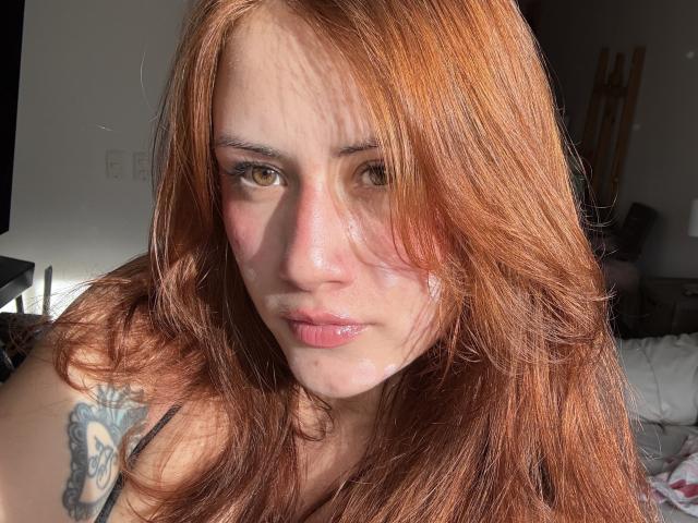 Malena Jones heeft rood haar, een tatoeage op haar linker schouder en een lichtrode huid met glanzende, roze lippen, en ze staart intens naar de camera..