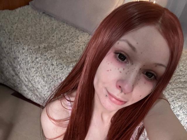 HannaCool has red hair, freckles, and a nose piercing. She's topless with visible cleavage, smiling at the camera. Her nose ring is on her left nostril.